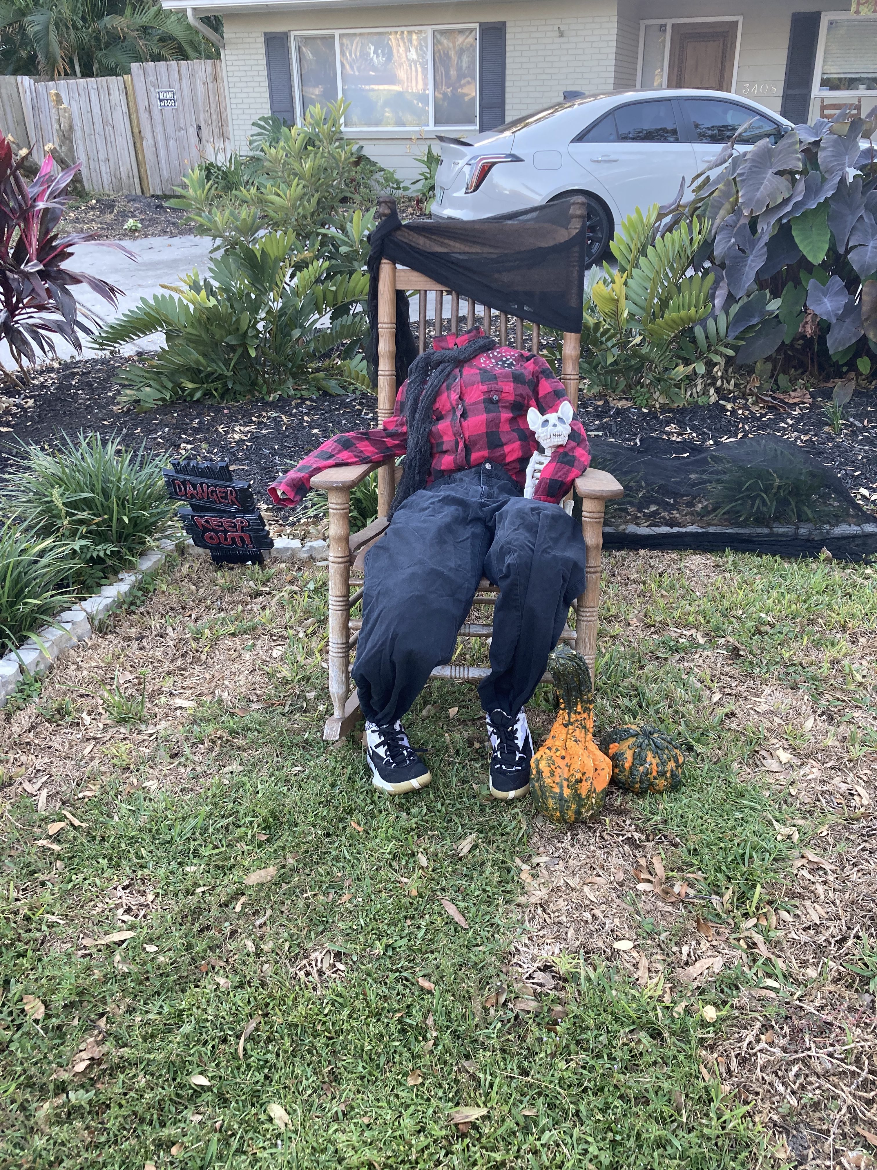 A life-sized scarecrow figure dressed in a red flannel shirt and black pants, sitting in a rocking chair on a front lawn, accompanied by a small toy. Nearby, there are ornamental gourds and a 'Danger Keep Out' sign.