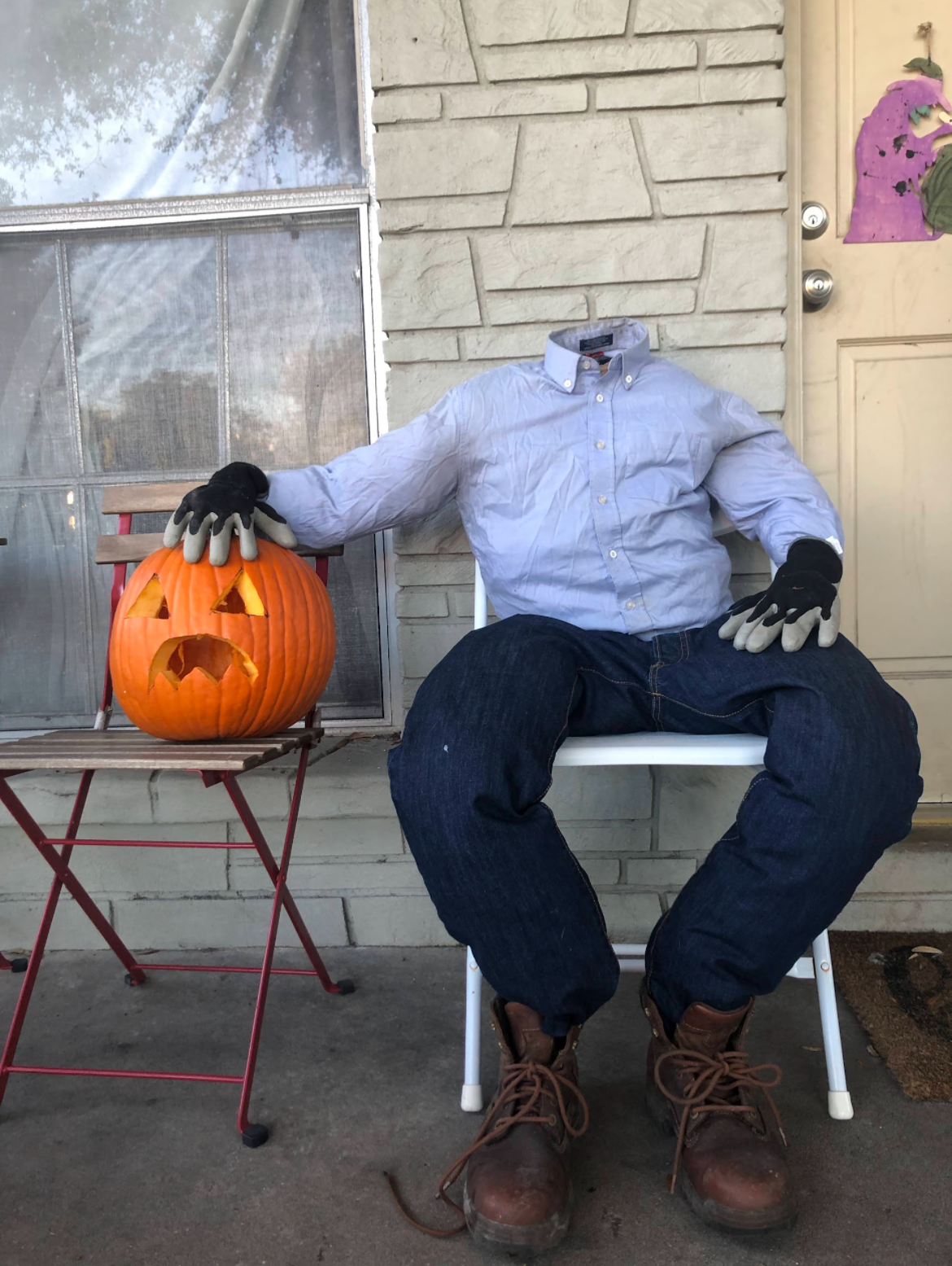 A headless scarecrow figure sitting on a chair next to a carved Halloween pumpkin with a surprised expression.