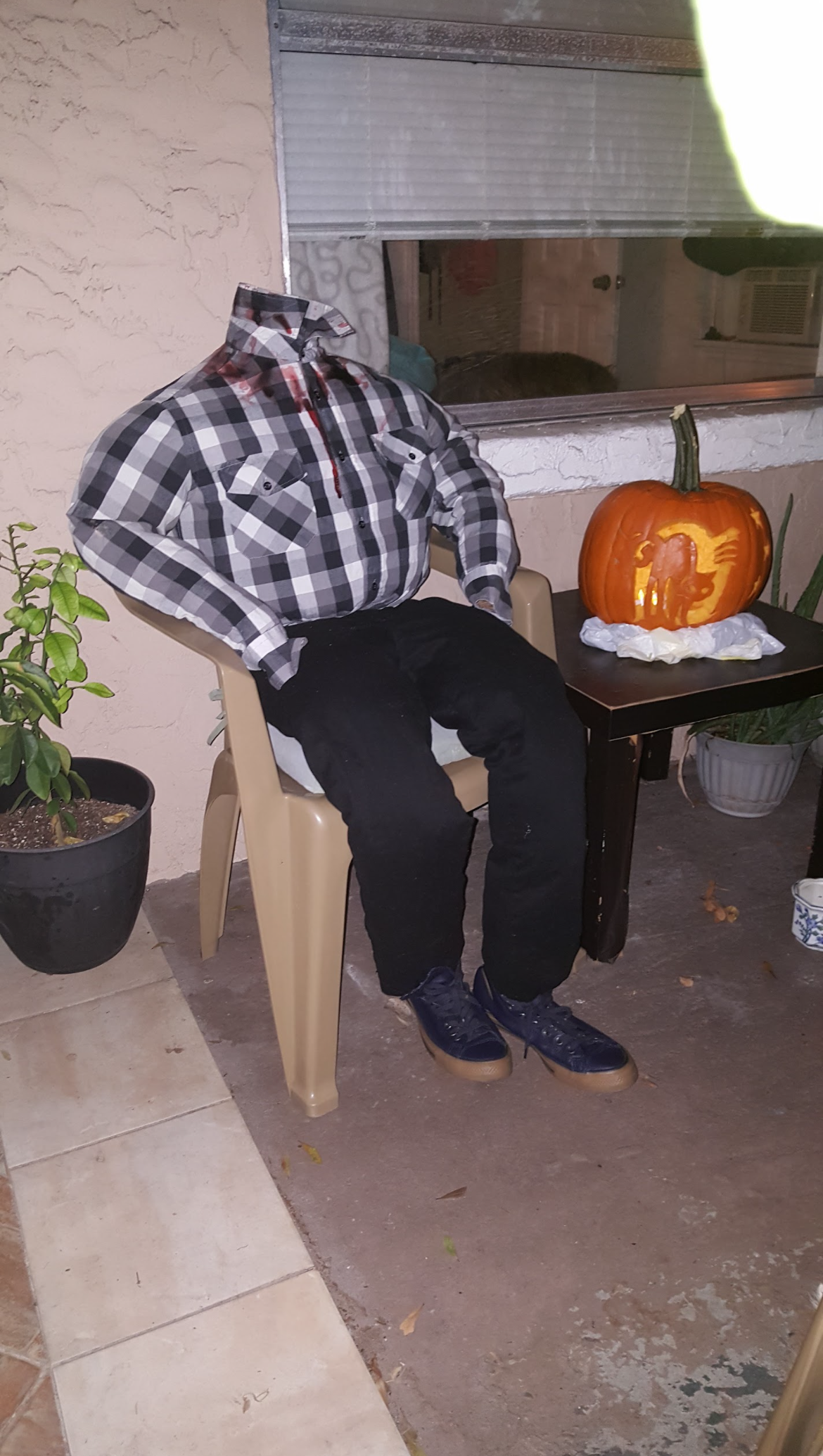 A headless mannequin wearing a plaid shirt and black pants sitting in a plastic chair, with a carved pumpkin on a nearby table.