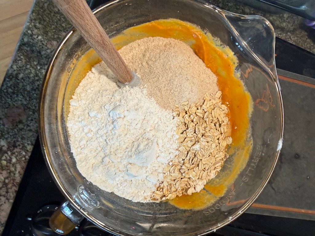 A mixing bowl containing flour, oats, and a wet mixture with a wooden spoon stirring the ingredients.