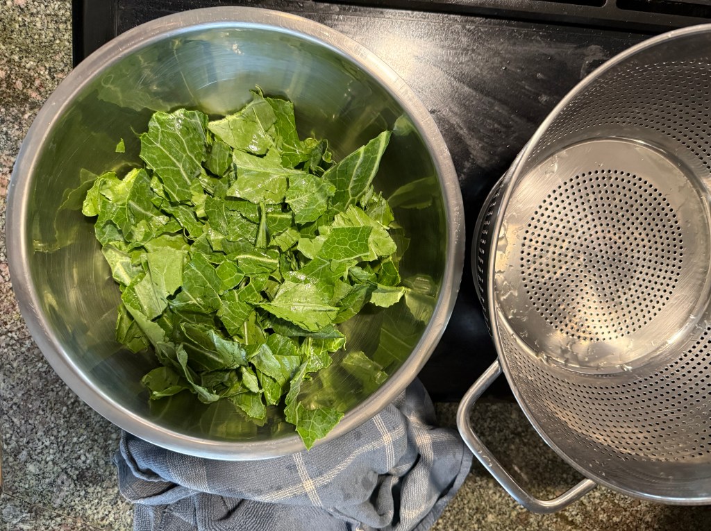 A stainless steel bowl filled with chopped leafy greens, next to a metal colander and a blue kitchen towel on a countertop.