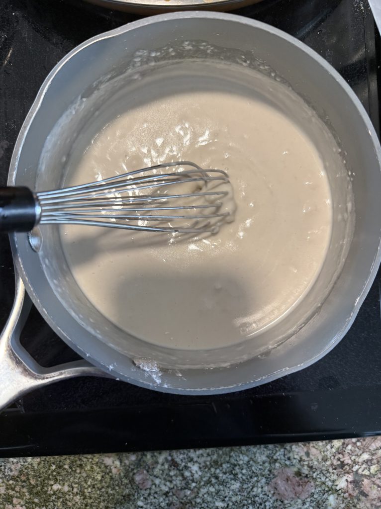 A saucepan with a smooth, pale batter mixture and a whisk inside, placed on a stovetop.